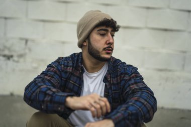 Ekose gömlekli ve kot pantolonlu genç bir adam sokakta oturur ve mesafeye bakar..