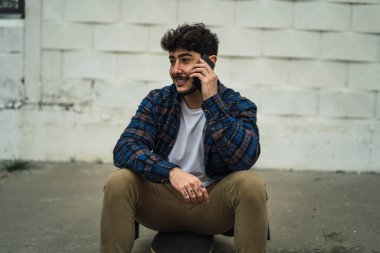 Cep telefonlu genç sakallı adam yerde oturuyor.