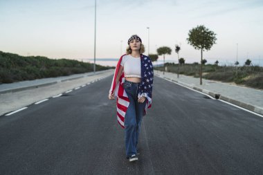 Amerikan bayrağı sallayarak ve el sallayarak Amerikan bayrağıyla yolda yürüyen gündelik kıyafetli genç bir kadın.
