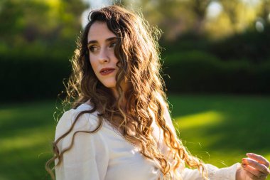 Beyaz bluzlu genç bir kadın yeşil bir parkta poz veriyor.