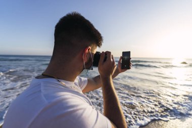 Plajda cep telefonuyla fotoğraf çeken sıradan giysili genç bir adam..