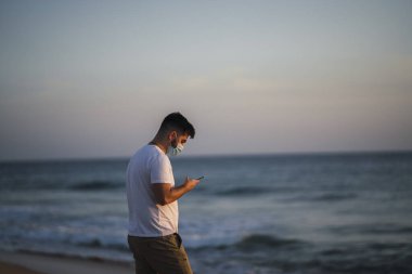 Genç adam gün batımında sahilde cep telefonu kullanıyor.