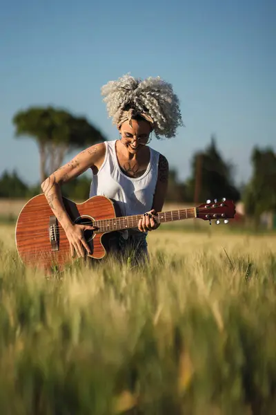 Yazlık elbiseli ve şapkalı bir kadın çayırda gitar çalıyor.