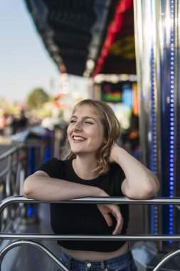 Genç bir kadın lunaparkta oturmuş kameraya bakıp gülümsüyor.