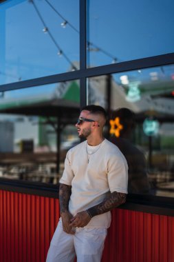 Sakallı, dövmeli, kırmızı tişörtlü ve beyaz gömlekli yakışıklı bir adam kahve dükkanında oturuyor..