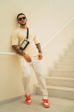 Beyaz tişörtlü, kot pantolonlu, metal bir merdivenin yanında poz veren şık bir adam..