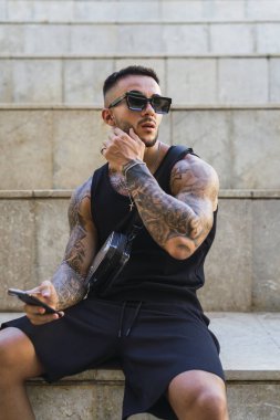 Dövmeleri olan yakışıklı Afrikalı genç adam bankta oturuyor.