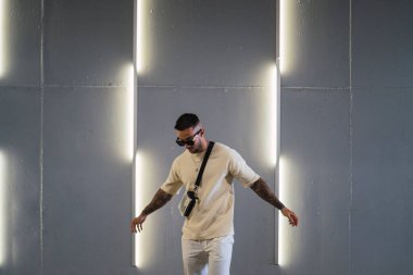 Güneş gözlüklü, sakallı, beyaz tişörtlü genç bir adam karanlık bir odada arka planda duruyor.