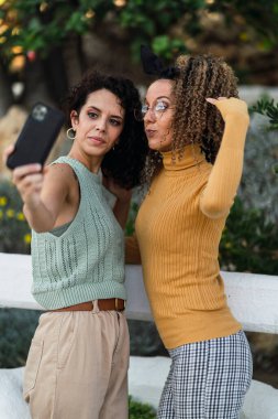 Kıvırcık saçlı iki güzel genç kadın sokakta selfie çekiyor..