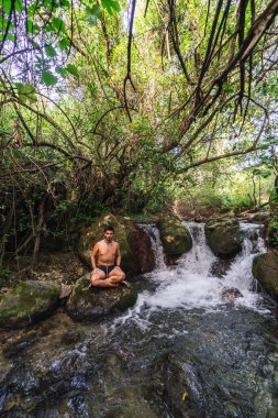 Siyah tişörtlü genç bir adam nehrin ortasındaki bir kayanın üzerinde oturuyor..