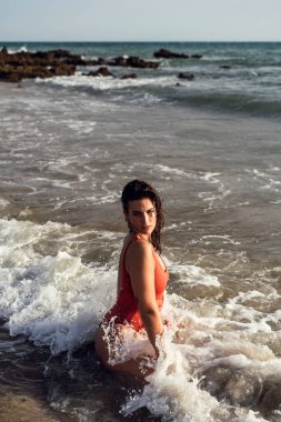 Genç bir kadın plajda kırmızı mayo ve beyaz elbiseyle poz veriyor.