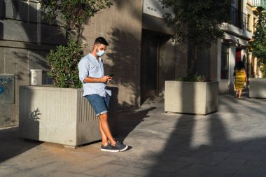 Koruyucu maskeli genç adam şehirde cep telefonuyla yürüyor.