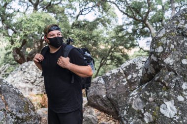 Sırt çantalı ve maskeli genç adam ormanda yürüyor.