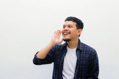Bağıran neşeli Asyalı adamın portresi. Günlük gömlekli mutlu genç erkek manken elleri ağızlarına yakın bir şekilde başka tarafa bakıyor, birini arıyor. Duyuru konsepti