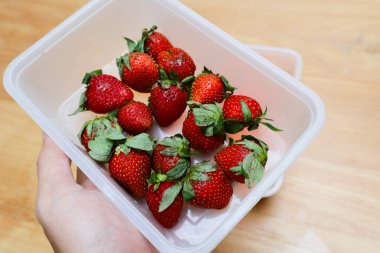 Tahta bir masada plastik bir kabın içinde çilekler.