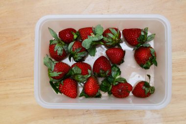 Tahta bir masada plastik bir kabın içinde çilekler.