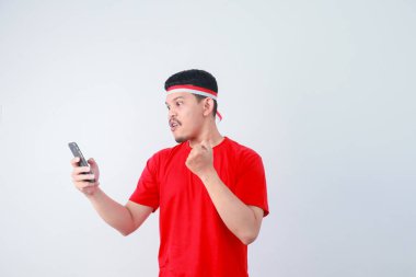 Excited young Asian man in casual t-shirt using mobile phone, getting good news isolated on grey background. indonesian independence day celebration concept