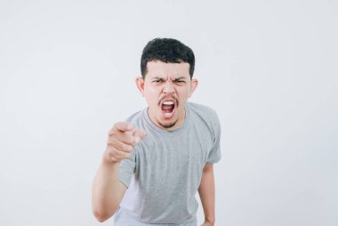 Gri tişörtlü genç Asyalı adam yoğun ve kızgın bir ifadeyle doğrudan kameraya bakıyor. Sade arka planda stüdyo çekimi. Günlük kıyafetlerini ve etkileyici görüntülerini yakalıyor.