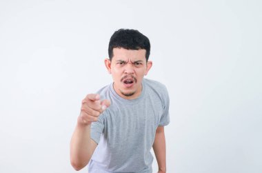 Gri tişörtlü genç Asyalı adam yoğun ve kızgın bir ifadeyle doğrudan kameraya bakıyor. Sade arka planda stüdyo çekimi. Günlük kıyafetlerini ve etkileyici görüntülerini yakalıyor.