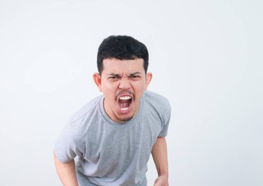 Gri tişörtlü genç Asyalı adam yoğun ve kızgın bir ifadeyle doğrudan kameraya bakıyor. Sade arka planda stüdyo çekimi. Günlük kıyafetlerini ve etkileyici görüntülerini yakalıyor.