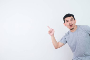 Gündelik tişörtlü, gri arka planda mutlu Asyalı bir adamın şaşırmış yüzü bir şeyi işaret ediyor. reklam kavramları