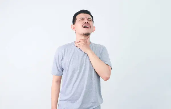 stock image A man in a grey shirt holds his throat with a pained expression, indicating a sore throat or discomfort. health and welness concept.