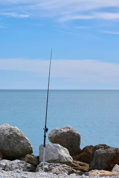 Single fishing rod on the stones of a breakwater in the sea. Blue sky with white clouds and calm sea