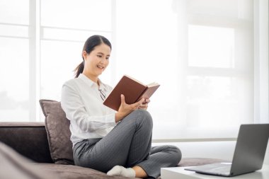 Evde oturan olgun bir kadın pencerenin önündeki modern koltukta oturuyor oturma odasında kitap okuyor ve evde dizüstü bilgisayarla çalışıyor.