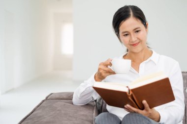 Evdeki oturma odasındaki kanepede kitap okuyan genç ve güzel bir kadın. Asyalı kadın tekstil kanepesinde oturmuş kahve içiyor. Beyaz duvar arkaplanı, kopyalama alanı, kapat.