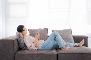 Mutlu olgun Asyalı kadın rahat koltukta rahat rahat oturup akıllı telefonuna mesaj atıyor, gülümseyen ev kadını cep telefonu kullanıyor, cihaz üzerinde kablosuz internet kullanıyor, evden online alışveriş yapıyor.