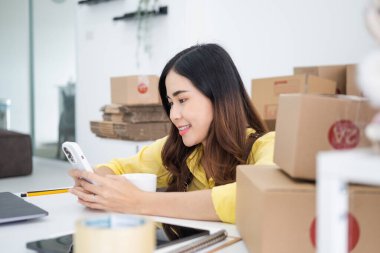 Gülümseyen mutlu Asyalı kadın cep telefonundan siparişleri kontrol ediyor, çevrimiçi bir mağazası var, özel bir nakliye şirketi aracılığıyla paketleyip gönderiyor. Çevrimiçi satış ve online alışveriş kavramları.