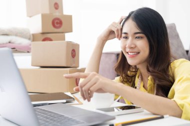 Asyalı iş kadını bilgisayar kullanır ve evde oturma odasında çalışır. Mutlu girişimci müşterinin siparişini kontrol ediyor. Parmak işaretini loptop 'a doğrult.