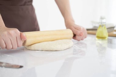 Önlüklü bir kadın beyaz masanın üzerinde yuvarlanıyor. Parlak, temiz bir mutfakta, ahşap bir oklavayla. Kapatın. Boşluğu Kopyala
