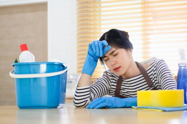 Mutfakta temizlik ürünleri ve ekipmanlarla oturan yorgun genç bir kadın. Ev işi konsepti.