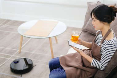 Robot elektrikli süpürge oturma odasını temizliyor. Genç bayan dinleniyor, evdeki kanepede oturuyor.