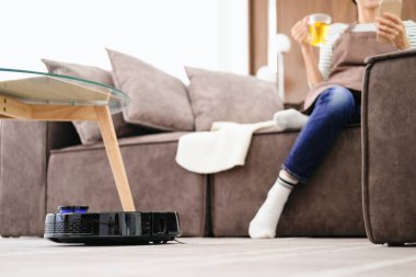Robot vacuum cleaner cleaning the living room. Young woman enjoy rest, sitting on sofa at home