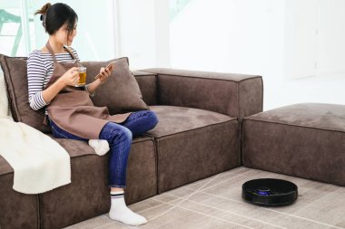 Robot vacuum cleaner cleaning the living room. Young woman enjoy rest, sitting on sofa at home