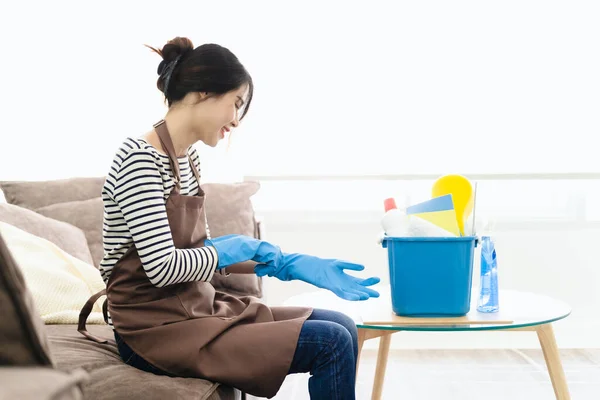 围裙上的女人戴上橡胶手套 坐在客厅里做家务活 有清洁设备的家庭主妇有乐趣 许多家务活 专业的清洁服务 — 图库照片
