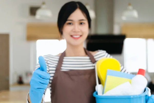 穿着棕色围裙和蓝色手套的年轻女性家庭主妇微笑着 手里拿着清洁剂瓶 露出大拇指 站在现代化的厨房里 提供专业的清洁服务 — 图库照片