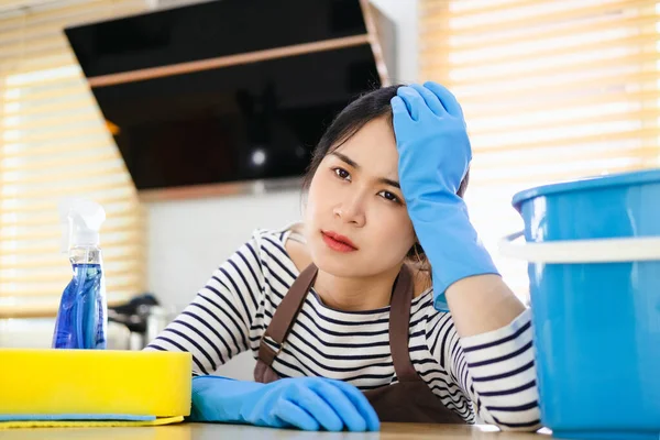 漂亮的亚洲家庭主妇做完家务活感到很疲倦 身穿棕色围裙和蓝色橡胶手套的精疲力竭的管家坐在厨房里 带着家用清洁用品 — 图库照片