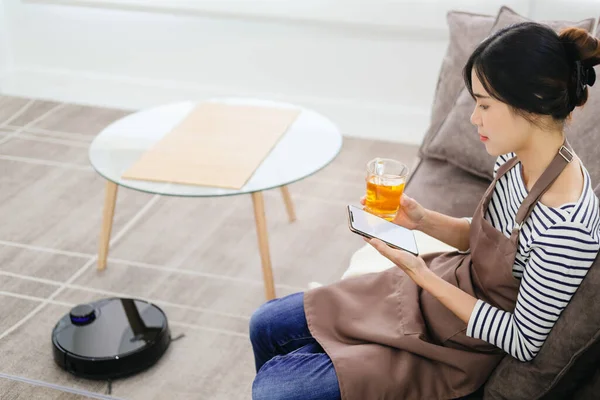 Robot vacuum cleaner cleaning the living room. Young woman enjoy rest, sitting on sofa at home