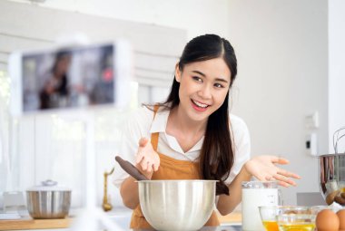 Blog yazarı pasta pişirme konusunda video dersi veriyor. Mutfakta önlüklü genç Asyalı kadın. Yayın çevrimiçi olduğunda, yazar yemek pişirme hakkında bir blog ya da kurs yönetiyor.