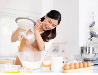 Modern mutfakta açık kahverengi önlük giyen genç bir kadın ekmek unu seçerken, karıştırmadan önce farklı malzemeler hazırlarken ve ev yapımı ekmek yapmak için fırında pişirmeden önce yoğururken eğleniyor..