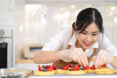 Genç ve mutlu ev hanımı elleri yemek eldivenleriyle yeni pişmiş turtayı yaban mersinli taze meyveyle süslüyor. Kadın aşçı önlük giyip meyveli tart yapıyor. Ev yapımı fırın..