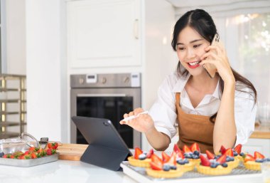 Fırın sahibi müşteriyle telefonda konuşuyor ve sipariş detaylarını yazıyor. Mutlu Asyalı fırıncının portresi kendi fırınında duruyor, tablet cihazını tıklatıyor ve akıllı telefondan konuşuyor..