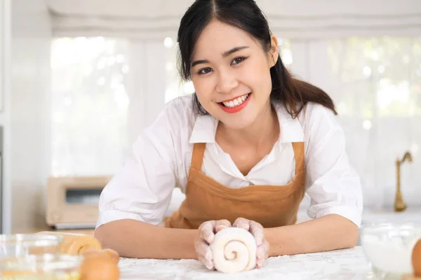 亚洲漂亮的成熟女人待在家里 花时间在厨房做饭 年轻迷人的女性穿着围裙 面带微笑 手里拿着酵母面团在桌上烘焙烘焙烘焙烘焙 在家里吃饭 — 图库照片