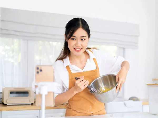 面包师在线课程 食物准备及烹调训练课程的概念 面带微笑的亚洲厨师在厨房里搅拌面粉 并用手机在三脚架上拍摄自己的视频 — 图库照片
