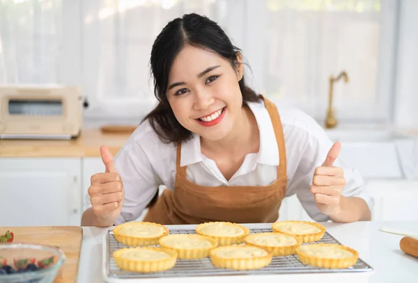 图为一位情绪激动的年轻亚洲女孩厨师在厨房里做菜时 大姆指翘起的照片 在明亮的厨房里 迷人的女人穿着围裙在白桌上烘烤新鲜水果馅饼 — 图库照片