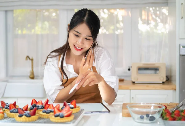 快乐的亚洲女面包师站在自己的面包店里 敲敲平板电脑 用智能手机说话的肖像 女工与客户在手机上交谈 烘焙房概念 — 图库照片