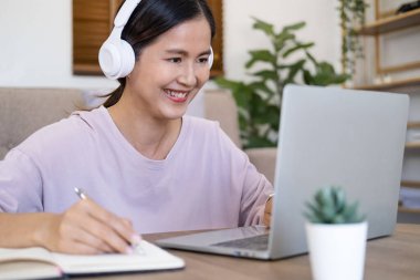 Mutlu genç bir kadın dil öğreniyor. Ders dinliyor, ders yazıyor. Dizüstü bilgisayara bakıyor, oturma odasında oturuyor, kız öğrenci Skype öğretmeniyle internetten kulaklık takıyor, uzaktan eğitim alıyor.,